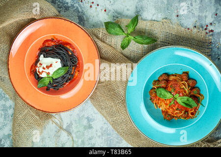 Blau Teller mit Nudeln und Fleisch Kugeln, orange Platte mit Tinte vom Tintenfisch Tinte. Graues Licht strukturierten Hintergrund. Schöne Servieren von Speisen. Restaurant Menü Stockfoto