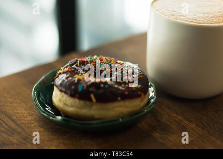 Kaffee mit einem gezeichneten Herz und Milch auf einen hölzernen Tisch in einem Café. Rosa Donut mit Streuung auf den Tisch neben der Kaffee Stockfoto
