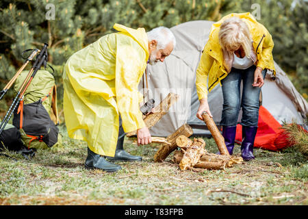 Senior Paar in gelbe Regenmäntel, Kamin auf dem Campingplatz in der Nähe von der Hütte in den Wald Stockfoto
