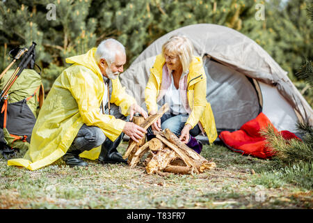 Senior Paar in gelbe Regenmäntel, Kamin auf dem Campingplatz in der Nähe von der Hütte in den Wald Stockfoto