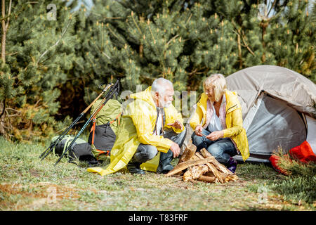 Senior Paar in gelbe Regenmäntel, Kamin auf dem Campingplatz in der Nähe von der Hütte in den Wald Stockfoto