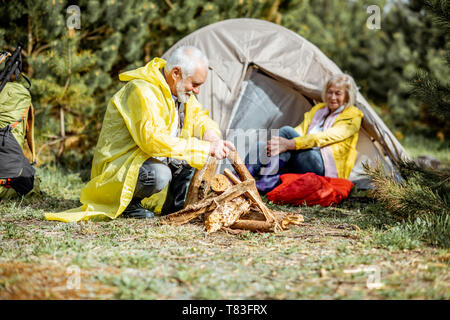 Senior Paar in gelbe Regenmäntel, Kamin auf dem Campingplatz in der Nähe von der Hütte in den Wald Stockfoto