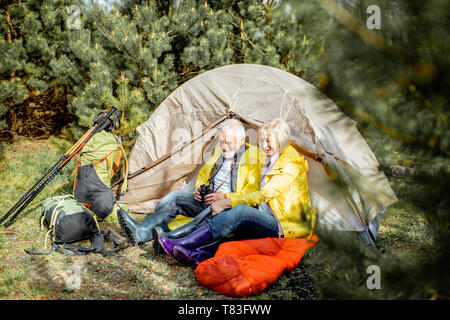 Senior Paar in gelbe Regenmäntel zusammen in der Nähe des Zeltes sitzen auf dem Campingplatz im Wald Stockfoto