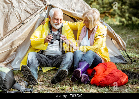 Senior Paar in gelbe Regenmäntel zusammen in der Nähe des Zeltes sitzen auf dem Campingplatz im Wald Stockfoto