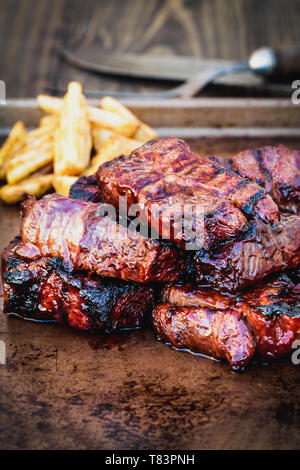 Bbq entbeintes Rindfleisch Rippen mit Barbecue Sauce und Kartoffelecken über eine rustikale Hintergrund. Extrem flache Tiefenschärfe mit unscharfen Hintergrund und Sele Stockfoto