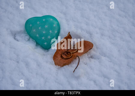 Zwei goldene Hochzeit Ringe in einem Herbstlich gefärbte Blätter, die an einem herzförmigen grünen Stein auf einem frischen Schnee Hintergrund. Close Up. Stockfoto