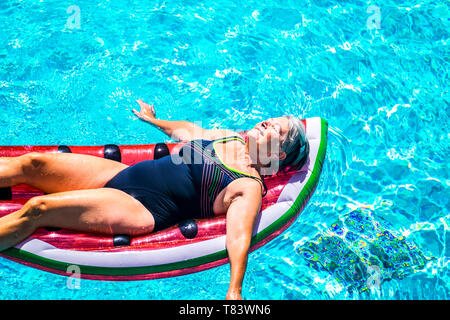 Gerne schöne Jahre älteren Dame genießen Sie die frische Wasser auf eine Wassermelone farbige lilo aufblasbare Matratze auf dem blauen Wasser des Pools lächelnd ein Stockfoto