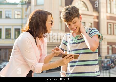 Eltern und Teenager, Beziehung. Die Mutter ihrem Sohn etwas im Handy zeigt, Junge ist peinlich, lächelnd, seine Hände auf seinen Kopf, Stadt St Stockfoto