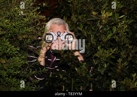 Ein grauhaariger Mann versteckt sich in Büschen mit einem Fernglas Spionage durch ein Fenster auf eine teilweise gekleidete Frau Stockfoto