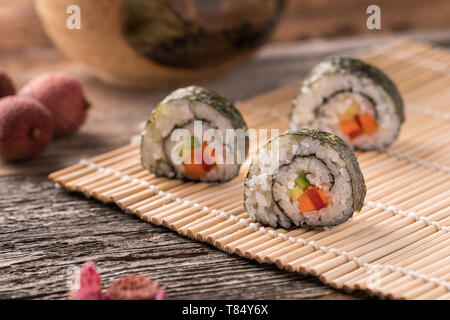 Drei Maki Sushi Rollen auf Bambus Matte auf Holz- Oberfläche, traditionellen asiatischen Tee-Schale, Litschi und Lychee Peel-close-up, gewinkelt, verschwommenen Hintergrund Stockfoto