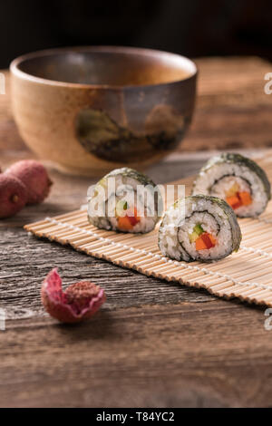 Drei Maki Sushi Rollen auf Bambus Matte auf Holztisch, traditionellen asiatischen Tee-Schale, Litschi und Litschis schälen - gewinkelt, unscharfen Hintergrund, vertikal Stockfoto