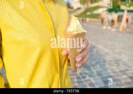 Nahaufnahme von gelb braun Zitrone Schokolade Eis in der Waffel Kegel in den Händen erwachsenen Frau, Frau im gelben Regenmantel outdoor, Frühling. Stockfoto