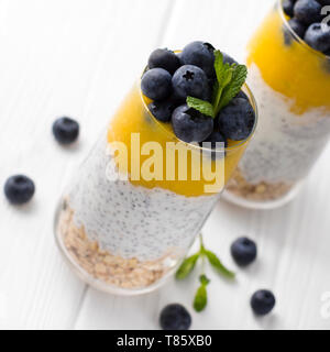 Glas Joghurt mit Chia Samen, Blaubeeren und Mango Mousse und Haferflocken auf einem weißen Holztisch. Stockfoto