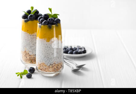 Glas Joghurt mit Chia Samen, Blaubeeren und Mango Mousse und Haferflocken auf einem weißen Holztisch. Stockfoto