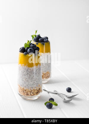 Glas Joghurt mit Chia Samen, Blaubeeren und Mango Mousse und Haferflocken auf einem weißen Holztisch. Stockfoto