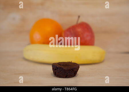 Früchte von Apfel, Orange und Banane mit einem kleinen Brownie im Vordergrund. Stockfoto