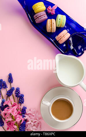 Tasse Latte, Makronen und Milch Glas auf Pastell rosa Hintergrund mit muscari und Hyazinthen Blumen dekoriert. Overhead shot, flach. Frühstück Konzept Stockfoto