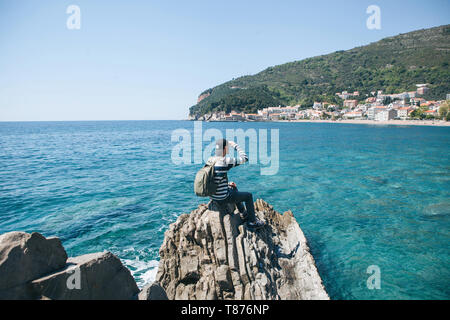 Ein Tourist mit einem Rucksack allein sitzt auf einem Felsen am Meer und in die Ferne schaut auf die Ansicht von Petrovac in Montenegro. Stockfoto