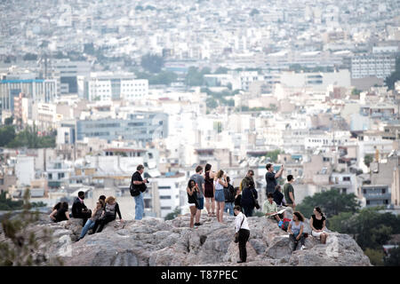 Touristen auf dem Akropolis-Hügel Athens City View Greece / ATHEN, Griechenland – Touristen versammeln sich auf den felsigen Hängen der Akropolis und genießen einen fantastischen Blick auf die weitläufige Stadt Athen darunter. Die antike Zitadelle, die zum UNESCO-Weltkulturerbe gehört, zieht jedes Jahr Millionen von Besuchern zu den Denkmälern aus dem 5. Jahrhundert v. Chr., darunter Parthenon und Erechtheion. Von diesem erhöhten Aussichtspunkt aus ist die gesamte Größe der modernen griechischen Hauptstadt sichtbar, die sich über die attische Ebene in Richtung der umliegenden Berge und des dahinter liegenden Meeres ausbreitet. Stockfoto