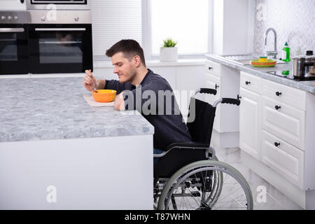 Seitenansicht der lächelnden behinderten Jungen Mann sitzt auf Rollstuhl trinken Suppe in der Küche Stockfoto