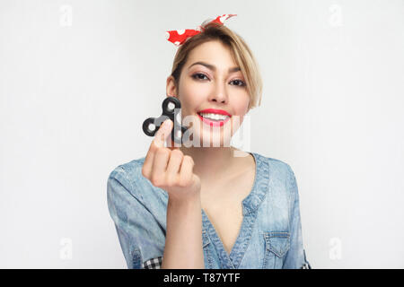 Portrait von schöne glückliche junge Frau in casual Blue Denim Shirt mit Make-up und roten Stirnband zu stehen und die Hand Spinner, toothy Lächeln. die Pflege von Stockfoto