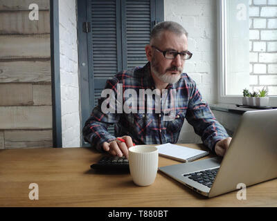 Ein männlicher Buchhalter arbeitet auf einem Computer zu Hause, zählt auf einen Rechner und macht sich Notizen in ein Notizbuch, ein freier Mitarbeiter bei der Arbeit, freier Platz für Text. Stockfoto