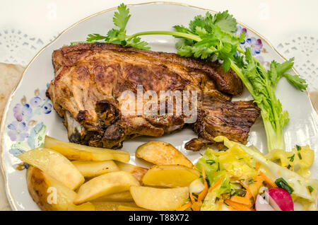 Großes Stück Schweinebraten und Bratkartoffeln mit Krautsalat, geschnittene Karotten mit Stücken von Rettich und einem Zweig Koriander auf einem porzellanteller Stockfoto