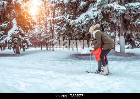 Eine junge Mutter unterstützt einen jungen Sohn, ein Junge von 3 Jahren. Im Winter draußen im Park, Hintergrund ist schneeverwehungen der Weihnachtsbaum. Freien Speicherplatz. Kinder- Stockfoto