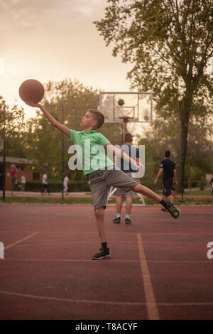 Ein 9 Jahre alter Junge, holding Basketball Ball in seiner Hand wie, wie es anzubeten. draußen auf einem Basketballplatz, mit den Menschen, die dahinter stehen. Stockfoto