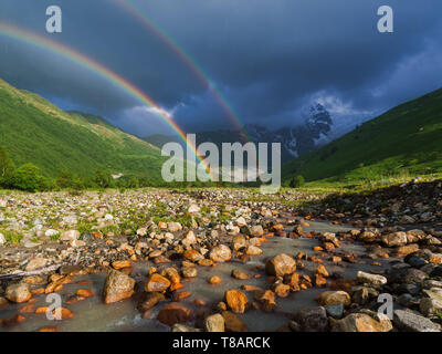 Regenbogen im Gebirge. Sommer Landschaft mit Fluss, Hügeln und einem stürmischen Himmel. Regen im Tal. Stockfoto