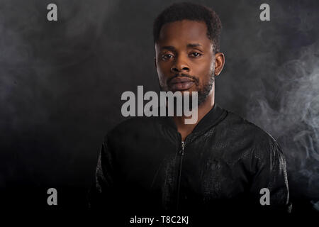 Headshot von schweren Dunkelhäutige männliche Holding Kinn geheimnisvoll Kamera suchen, in schwarz windjacke Jacke, wristwatces gekleidet, gegen die ständigen Stockfoto