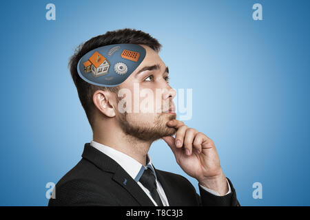 Junge Unternehmer denken mit weißen Haus, Zahnräder, Ziegel, Schraubenfedern, in seinem Kopf auf blauem Hintergrund Stockfoto