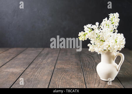 Weißer Keramik Vase mit weißen Flieder Blumen auf Holztisch. Wellness im Frühling oder im Sommer zur Aromatherapie Konzept. Mock up. Kopieren Sie Platz. Dark Photo Stockfoto