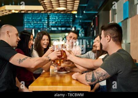 Junge weibliche und männliche Freunde halten Pints mit Bier und Toasten in der Kneipe. Gerne Gesellschaft bei Tisch sitzen, an einander suchen, Lachen und Reden im Cafe. Konzept der Brauerei und Spaß. Stockfoto