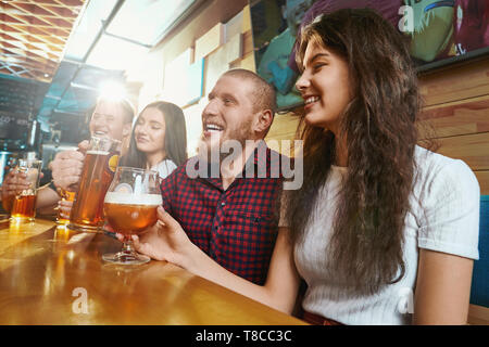 Seitenansicht des Fröhlichen Freunden Lachen und Scherzen beim Trinken von Bier im Pub. Glückliche Männer und eine Frau sitzen gemeinsam an der Bar und Toasten am Wochenende. Konzept von Glück und Spaß. Stockfoto