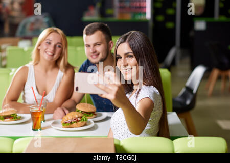 Schöne fröhliches Mädchen, Telefon und unter selfie mit Freunden gegenüber sitzt im Cafe. Lächelnd Unternehmen am Telefon und posieren beim Stillstehen. Konzept der Essen und lecker Essen. Stockfoto