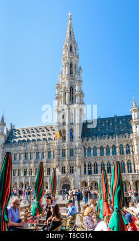 Touristen mit einem Drink auf der Terrasse eines Cafés auf dem Grand Place in Brüssel, Belgien, gegenüber dem Rathaus und der 96 Meter hohen Glockenturm. Stockfoto