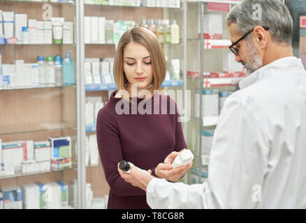 Reifen Apotheker bietet dem Kunden medizinische Produkte in Drogerie. Schöne Frau mit weißen kosmetische Flaschen, wählen. Chemiker in graue Haare, Brille und weißen Kittel. Stockfoto