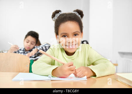 Schön, schöne Mädchen, Schulkind lächeln, sitzen am Schreibtisch, Bleistift, Schreiben in copybook, an der Kamera schaut. Fröhlicher, lustiger Schülerin im Unterricht in der Lektion in der Schule stellen. Stockfoto