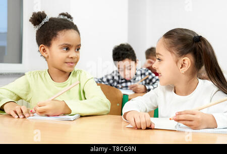 Nette, hübsche Schülerinnen am Schreibtisch im Klassenzimmer sitzen, Halten in der Hand pen Il, an einander suchen, posieren. Schüler der Grundschule schreiben in Copybook auf Lektion. Stockfoto
