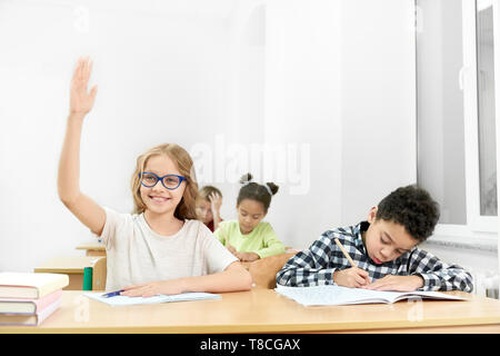 Schulkinder, Jugendliche am Schreibtisch im Klassenzimmer sitzen. Schöne, glückliche Schülerin in Blau Brille mit erhobener Hand an der Kamera. Konzentrierte junge Schreiben mit Stift in Copybook. Stockfoto