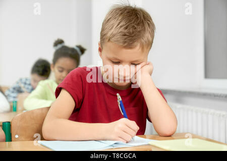 Primäre Schüler am Schreibtisch sitzen, unten schauend, schiefen Kopf auf die Hand und Schreiben mit blauen Stift in Copybook im Klassenzimmer. Portrait von jungen, fleißigen Schüler in der Klasse. Stockfoto