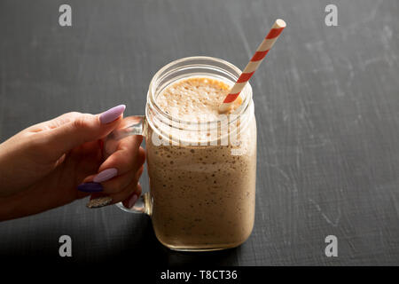 Woman's Hand mason Glas Glas Becher gefüllt mit Banane, Kiwi, Apfel Smoothie über schwarze Oberfläche, Low Angle View. Close-up. Stockfoto