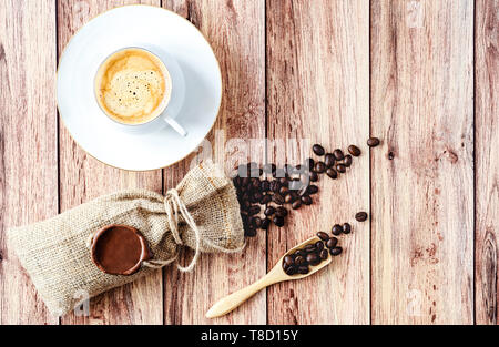 Blick von oben auf eine Tasse heißen Kaffee und Kaffeebohnen in eine hölzerne Schaufel und Auslaufen aus einem hessischen Beutel auf Holz- rustikalen Tisch. Platz für Text. Stockfoto