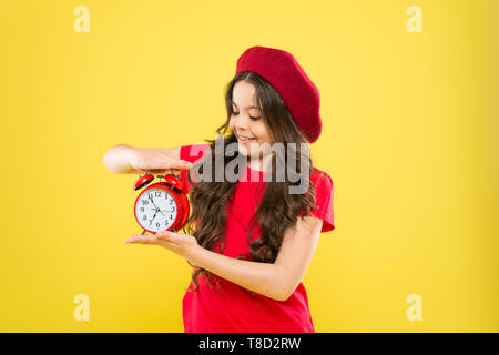 Glückliches Mädchen mit langen Locken in Baskenmütze. Kind mit Wecker. Zeitlose Mode. Pariser Kind auf Gelb. Schönheit Friseur. Kleines Mädchen im französischen Stil hat. Mädchen zeigt die Zeit auf der Uhr. Stockfoto