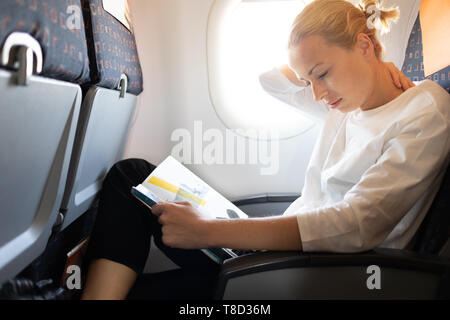 Frau Gefühl Nackenschmerzen beim Lesen im Flug Magazin auf langen interkontinentalen Flug. Weibliche Reisende unbequem im Pkw sitzen Stockfoto