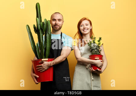 Gerne fröhliche junge Paar mit Blumen an die Kamera posieren. Isolierte gelben Hintergrund, studio Shot, Beruf, Hobby, Emotion, Gefühl Stockfoto