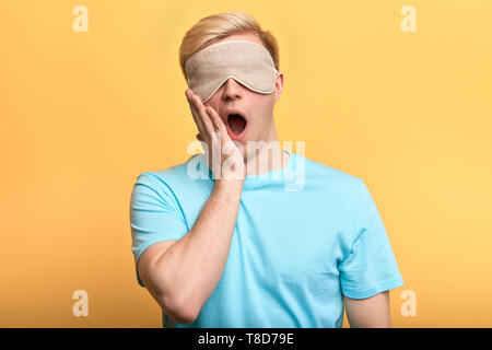 Junger Mann leger in Schlafmaske mit plam auf seinem Mund gähnend am Morgen angezogen. Foto in der Nähe. Stockfoto