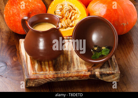 Kürbissuppe serviert in einem Tonkrug/Topf, serviert auf einem Holzbrett mit ganzen Kürbissen im Hintergrund. Stockfoto