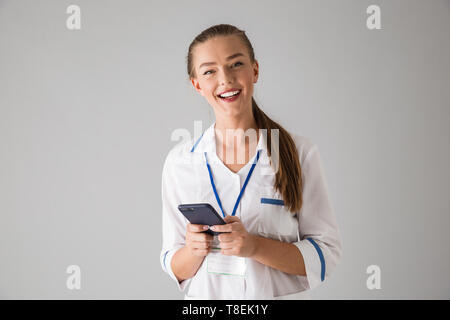 Foto von Eine schöne glückliche junge Frau cosmetologist Arzt über graue Wand Hintergrund holding Handy getrennt durch. Stockfoto